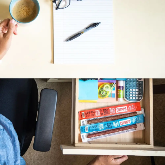A desk with coffee, paper, pen, and an open drawer containing snacks and office supplies.