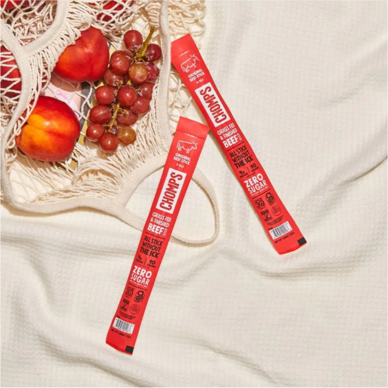 Two beef snack sticks on a textured surface beside a bag of peaches and grapes.