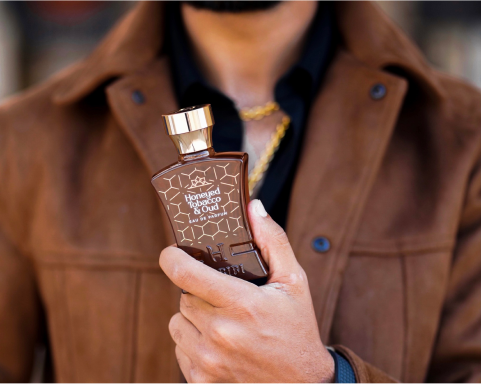 A person holding a brown bottle of Honeyed Tobacco & Oud perfume, wearing a brown jacket.