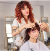 Hairstylist with curly hair styling a seated client's hair in a salon.