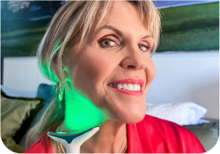 Person using a glowing green skincare device on their neck, smiling in a cozy setting.