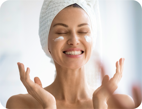 Smiling woman applying face cream with towel wrapped around her head.