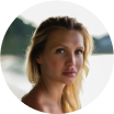 A woman with blonde hair standing near a blurred natural background.