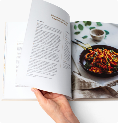 A person holding an open cookbook showing a pasta dish recipe.