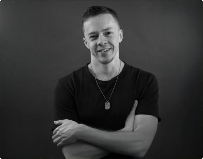 A person smiling with arms crossed, wearing a dark shirt and necklace.