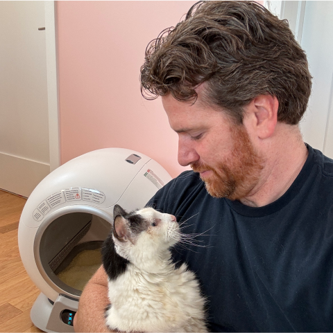 Man holding a cat in front of an automatic litter box.