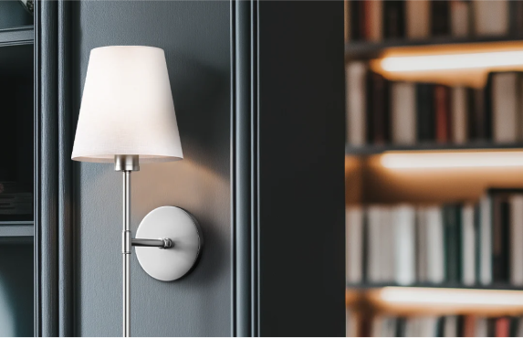 Wall-mounted lamp with a white shade beside a blurred bookshelf background.