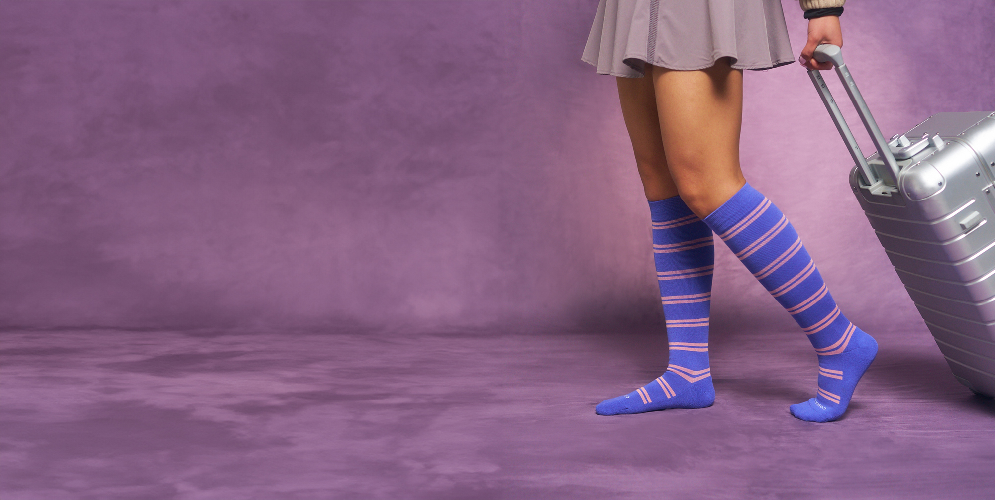 Person in striped socks pulling a silver suitcase on a purple background.