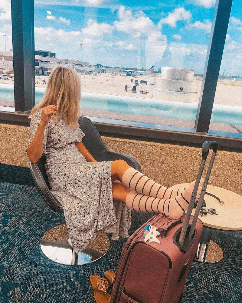 Person sitting in airport lounge with suitcase, looking out at runway.