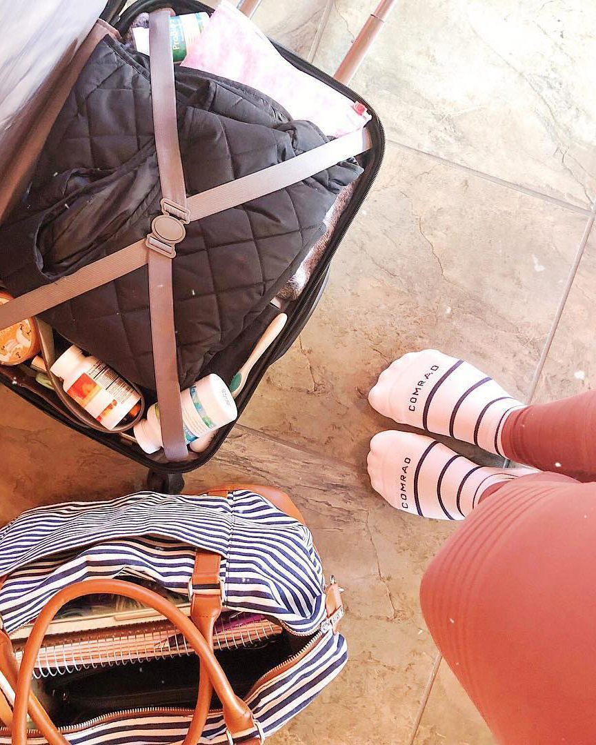 Open suitcase with clothes and toiletries, striped bag, and feet in socks on tiled floor.
