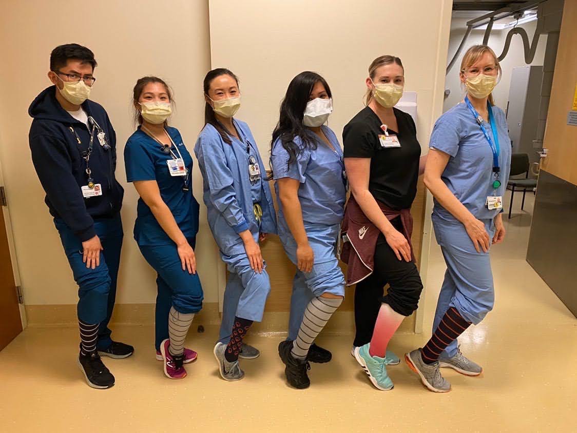 Six healthcare workers in scrubs and masks showing off colorful socks.