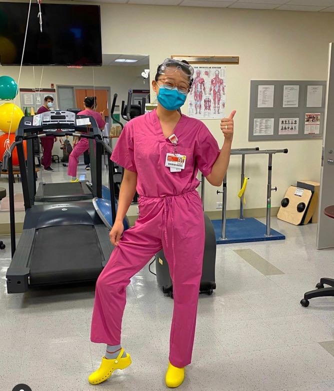 Person in pink scrubs and yellow shoes giving a thumbs up in a medical setting.