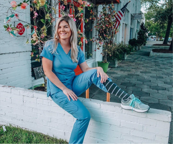 Nurse in blue scrubs sitting on a white wall, wearing striped Comrad compression socks and sneakers
