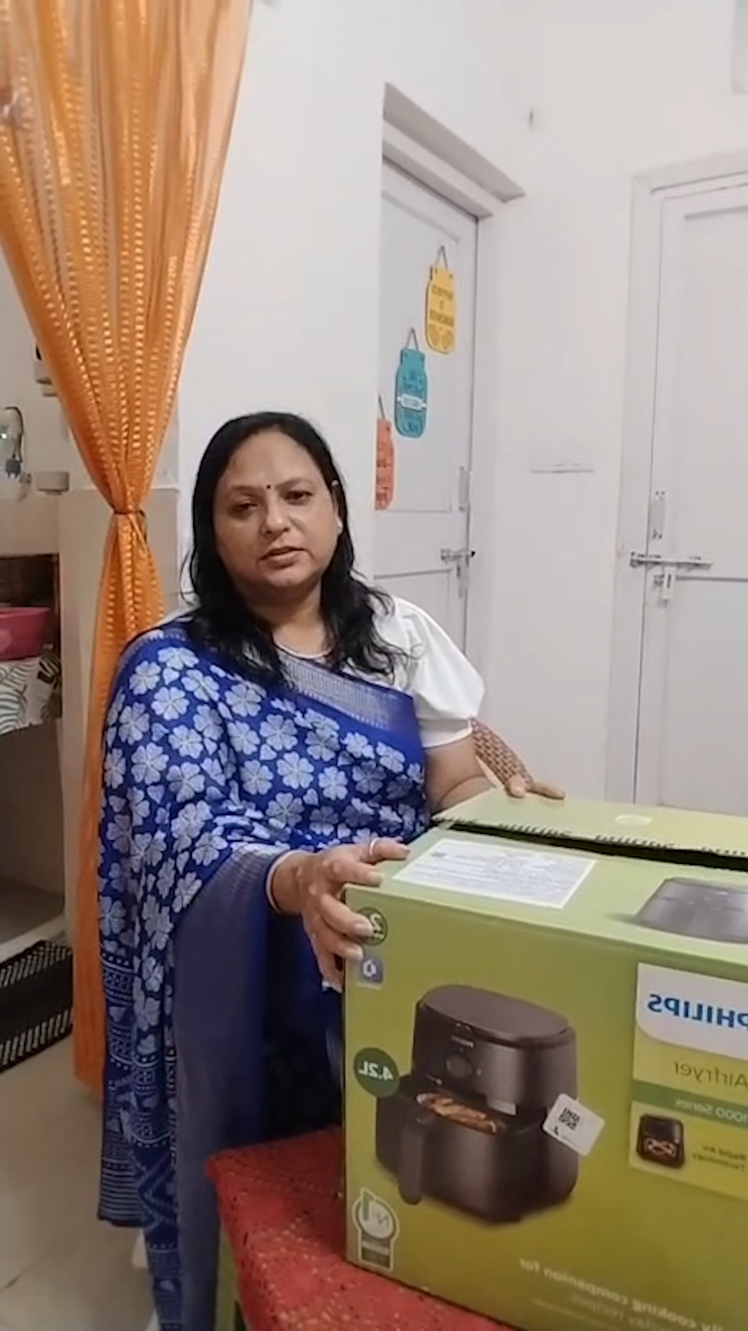 A woman in a blue sari stands behind a large box for a Philips air fryer.