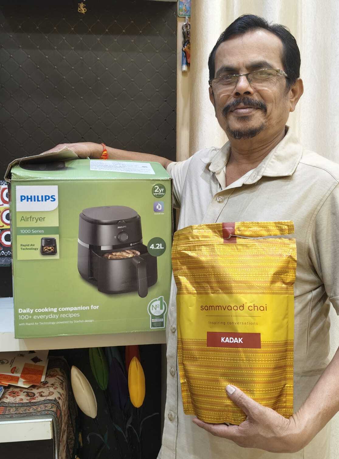A man holds a bag of Sammvaad Chai tea and stands beside a Philips Airfryer box.