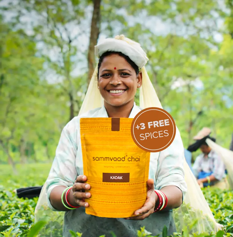 A smiling woman in a tea field holds up a yellow pouch of Sammvaad' Chai.