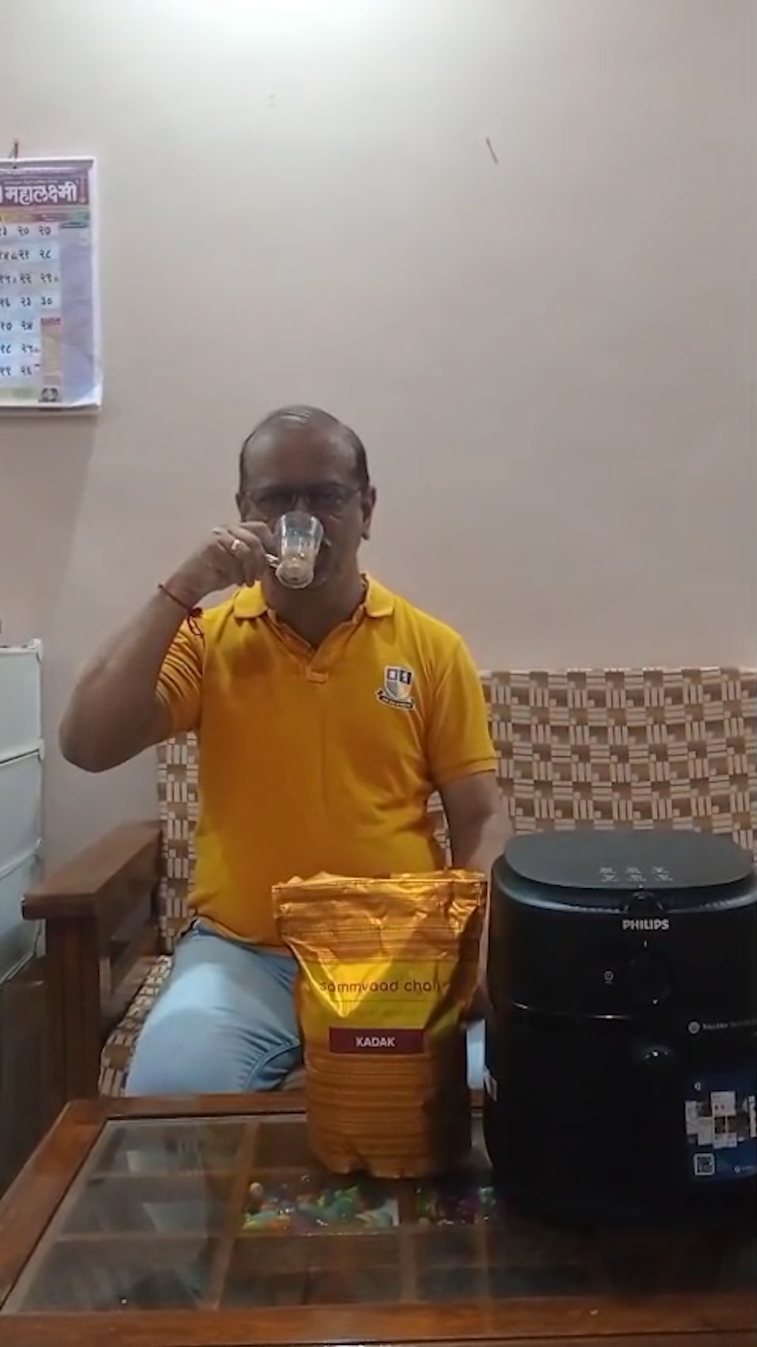 A man in a yellow shirt sips from a glass while sitting on a couch in a living room.