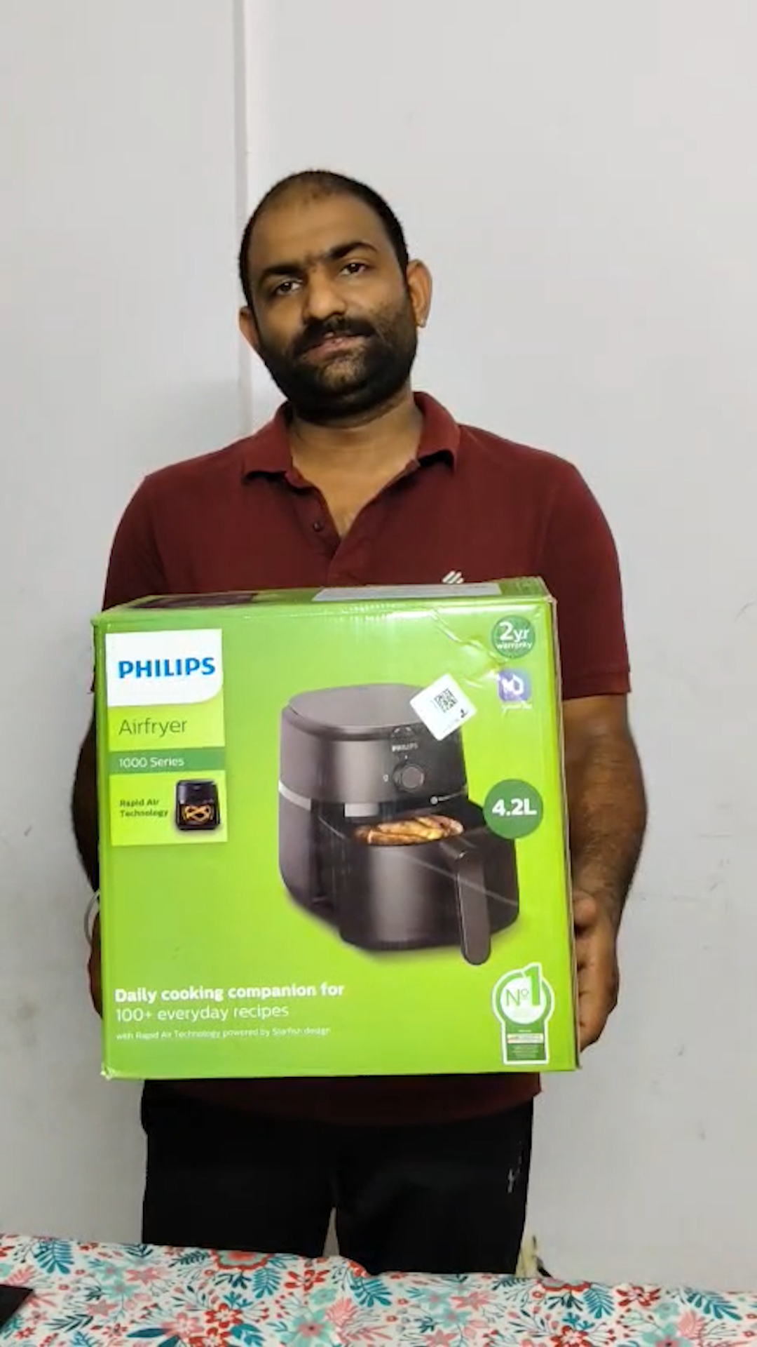 A man in a maroon polo shirt holds up a large green box for a Philips Airfryer.