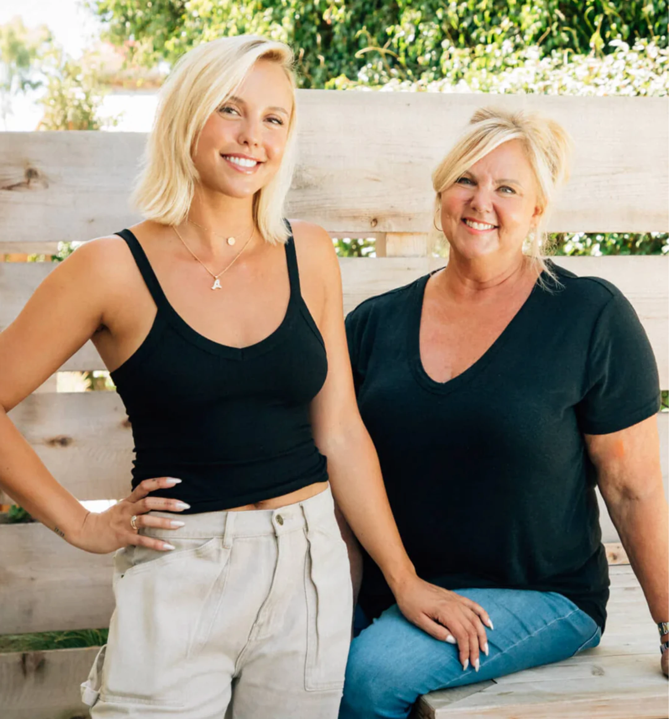 Two women smiling outdoors, standing and sitting on a wooden bench.