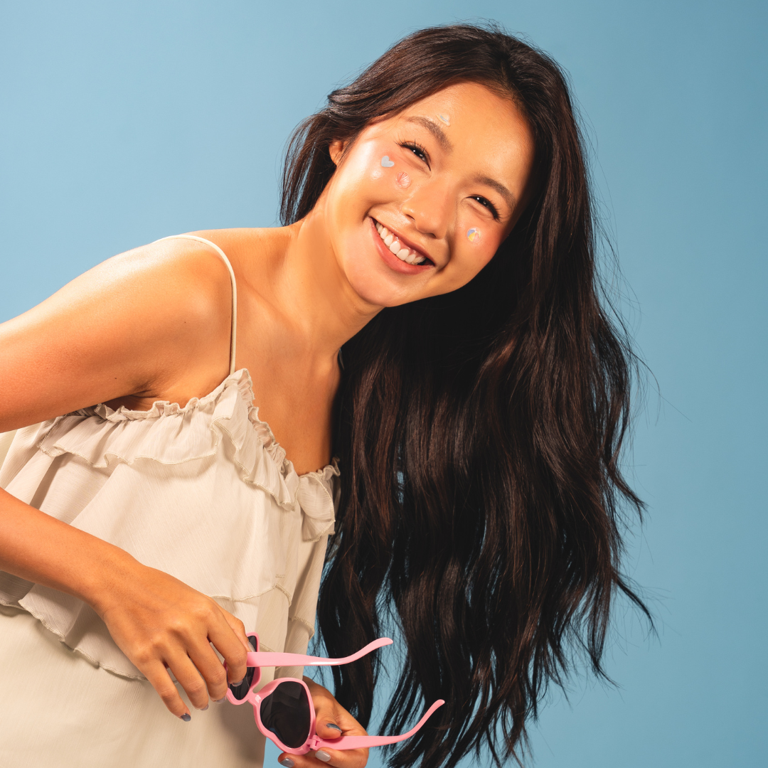 A smiling woman with heart-shaped stickers on her face holds a pair of pink heart-shaped sunglasses.