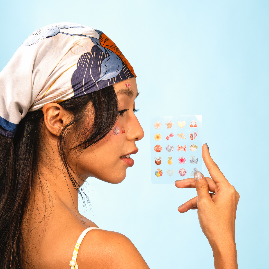 Profile of a woman in a headscarf holding a sheet of beach-themed stickers, with some on her face.