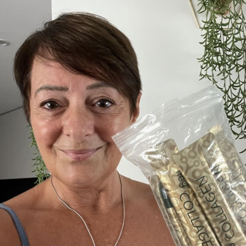 A smiling woman with short dark hair holds up a clear bag filled with gold collagen packets.