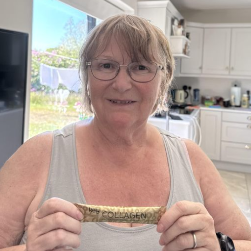 A woman with glasses smiles while holding a single-serving packet of key COLLAGEN in a kitchen.
