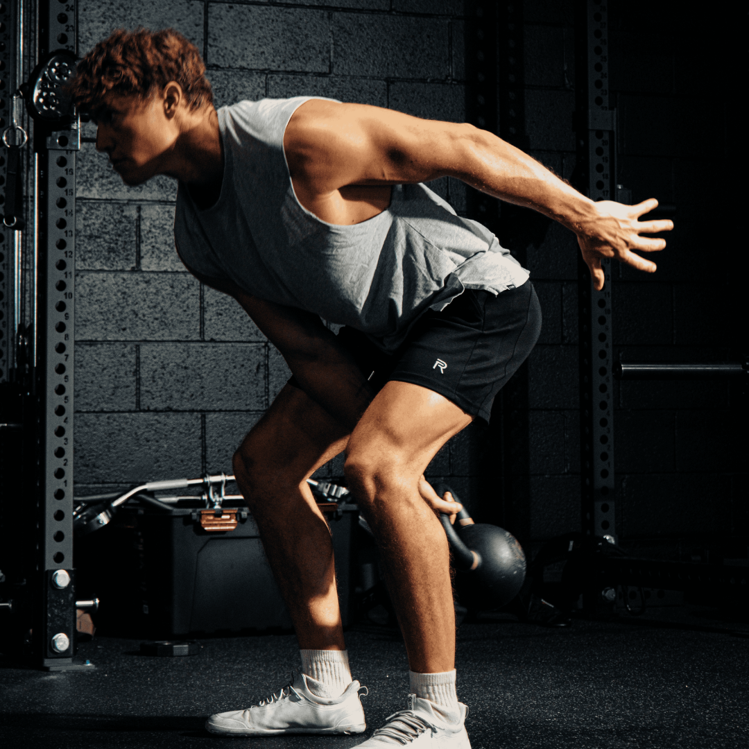 A man in a grey tank top and black shorts doing a kettlebell swing in a gym.