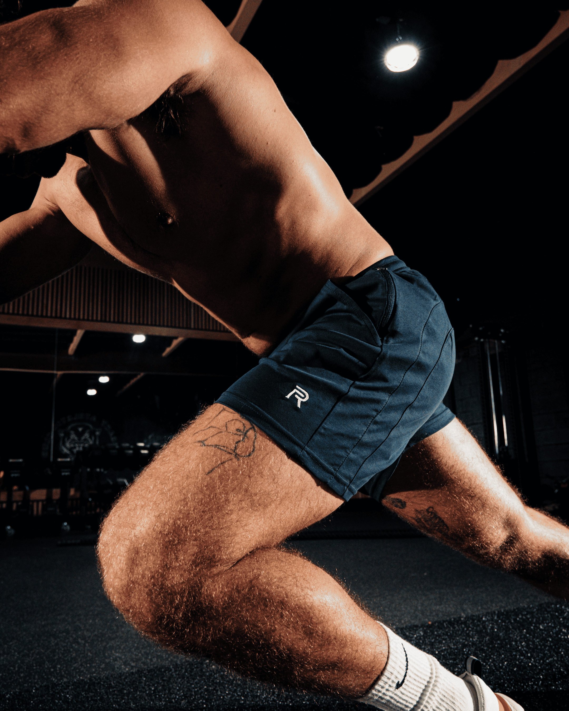 A close-up of a muscular, shirtless man in motion wearing navy athletic shorts in a dimly lit gym.