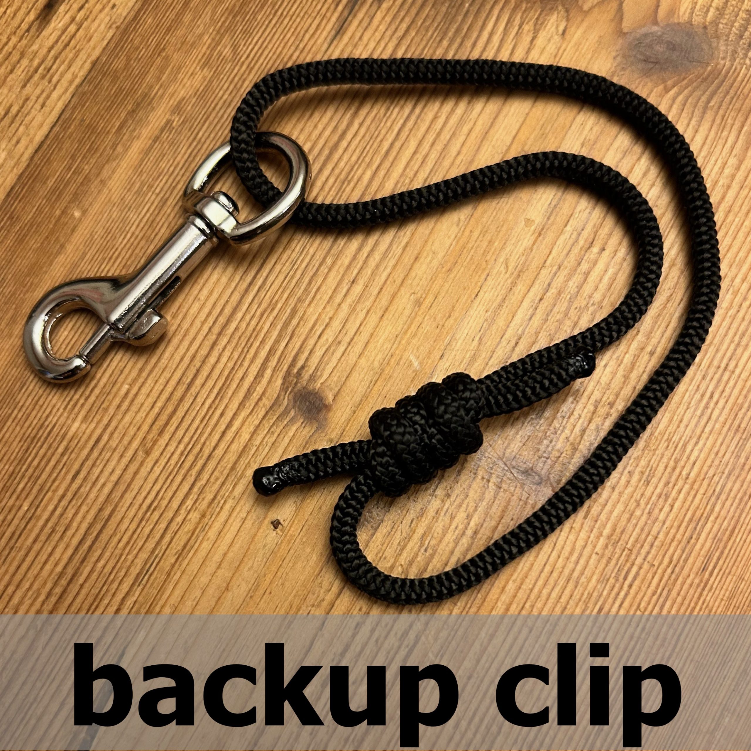 Black rope with a metal clip on a wooden surface, labeled 'backup clip'.