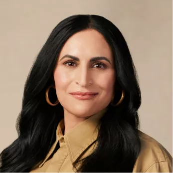 A headshot of a woman with long dark hair, wearing a tan collared shirt and gold hoop earrings.