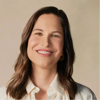 A headshot of a smiling woman with long brown hair, wearing a white collared shirt.