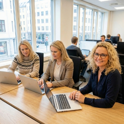 Three women work on laptops at a conference table in a modern office, with other colleagues behind them.
