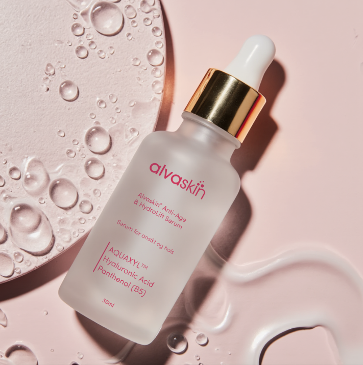 Frosted glass bottle of skincare serum with gold dropper on a pink surface with water droplets.