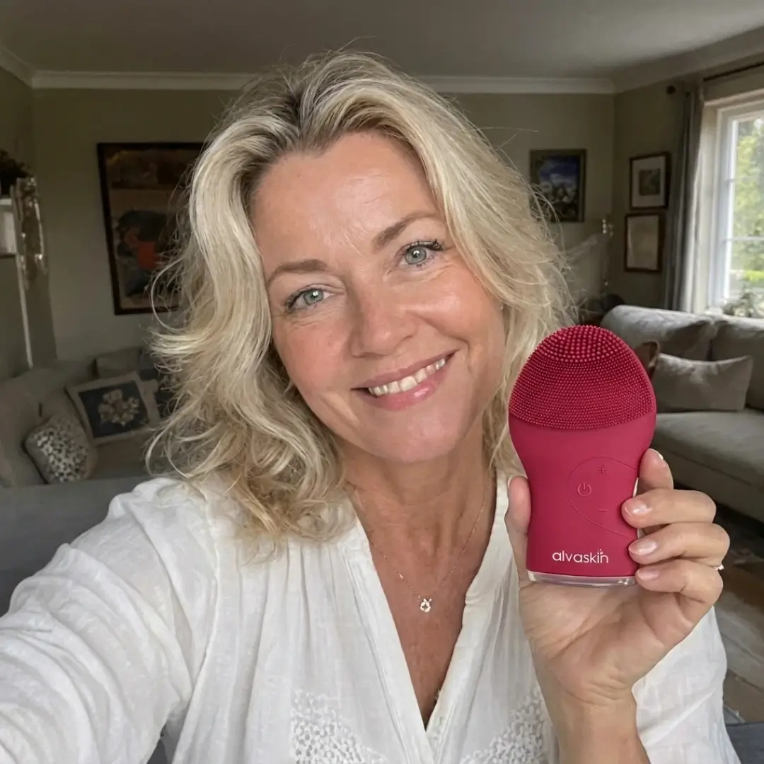 A smiling blonde woman holds up a red Alvaskin electronic facial cleansing brush in a living room.