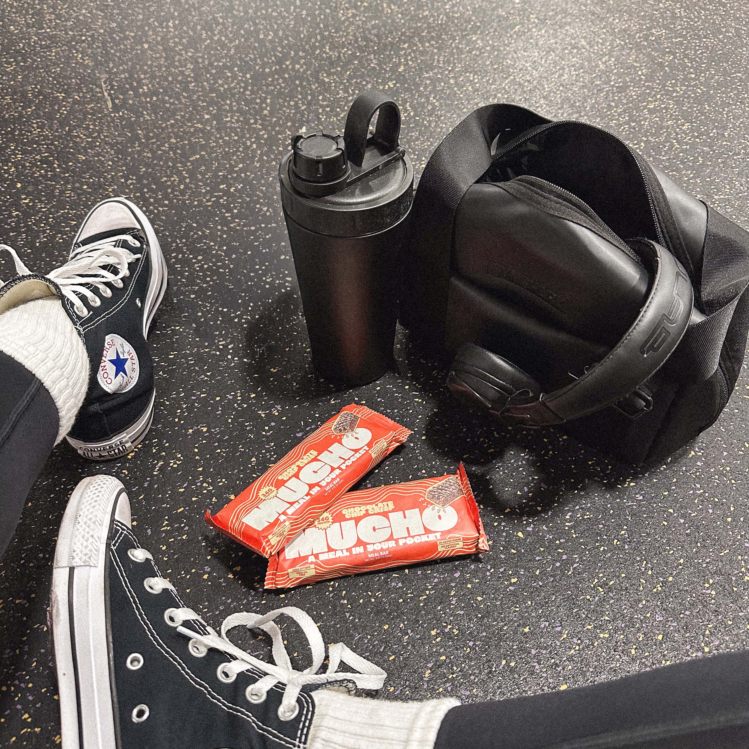 Gym bag, protein bars, shaker bottle, and sneakers on speckled floor.