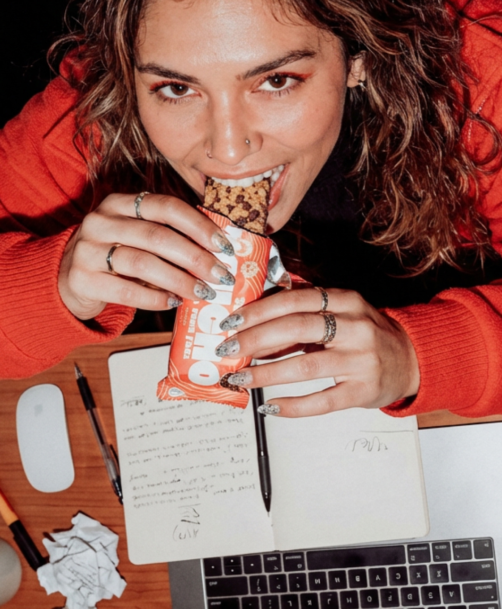 A woman in a red sweater eats a snack bar while working at a desk with a laptop.