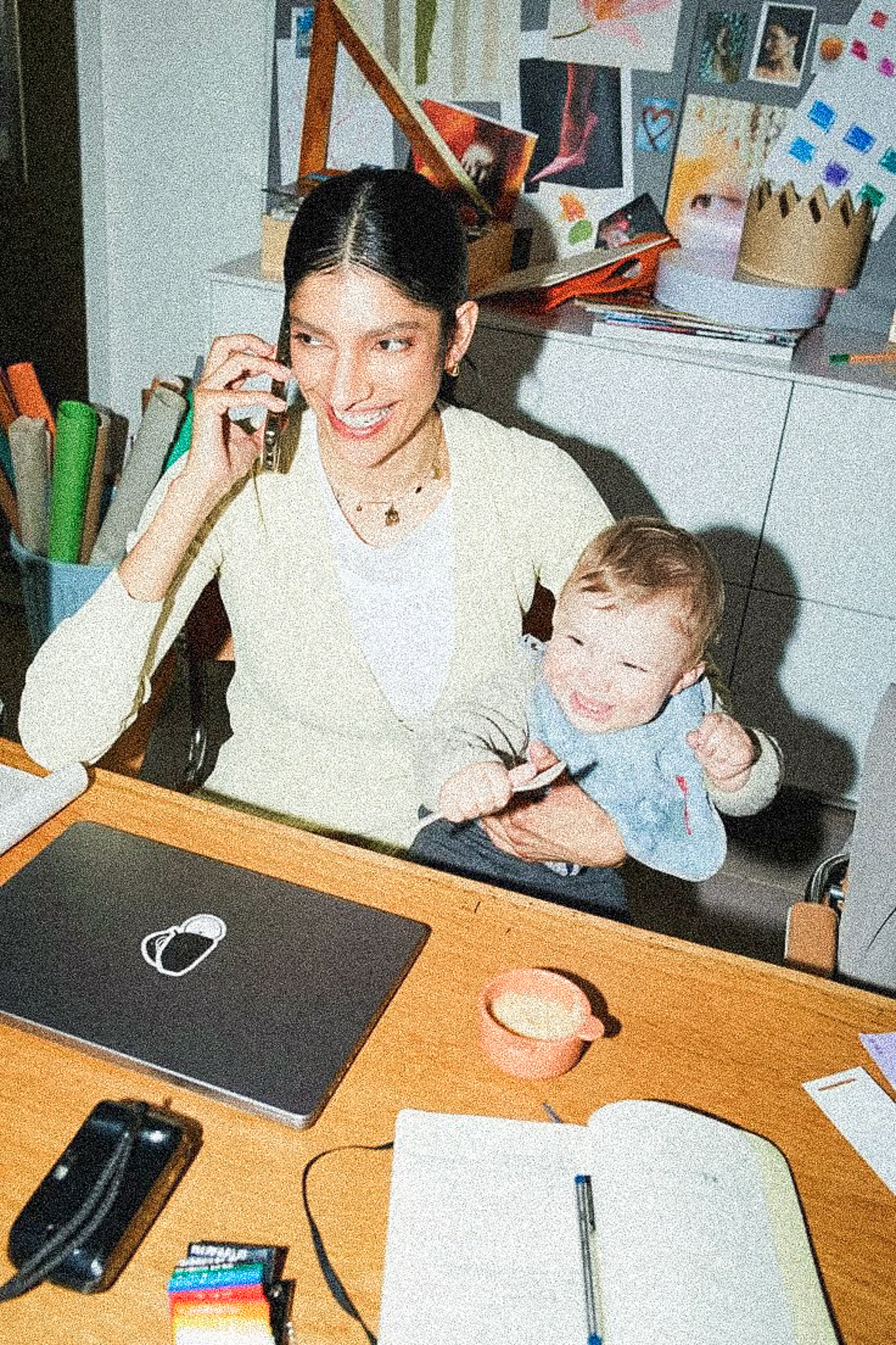 Person on phone with child on lap, desk with laptop and notepad.