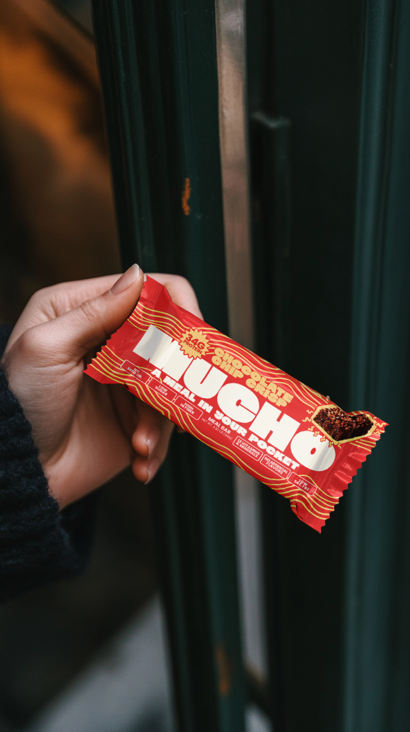 A hand holds a red-wrapped Mucho chocolate chip crisp meal bar in front of a dark green door.