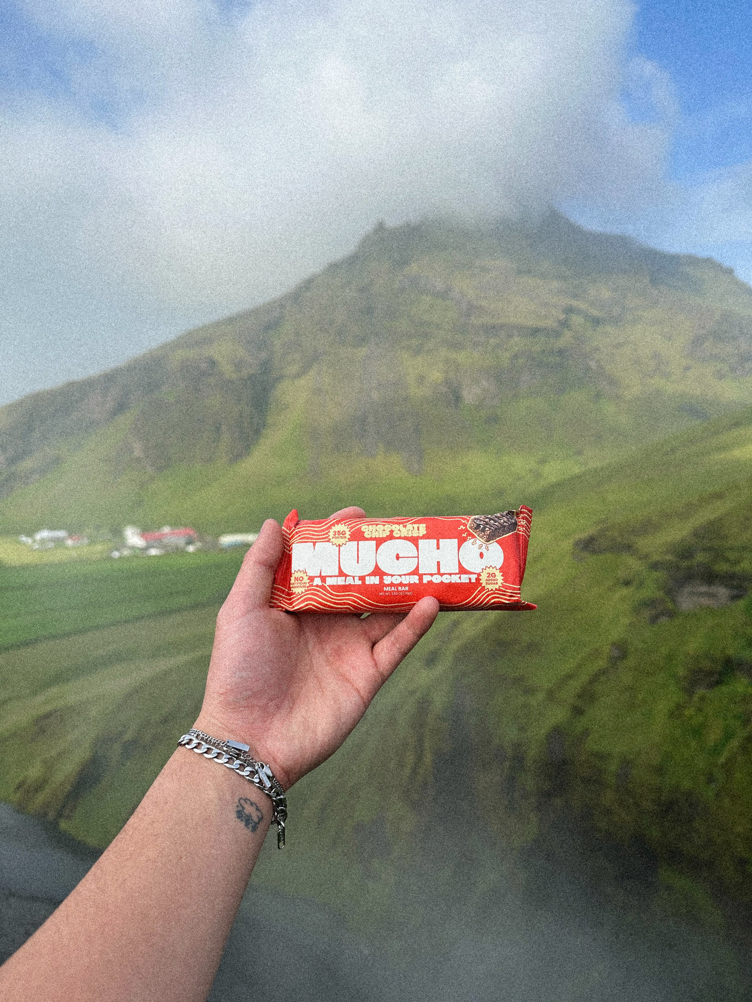 Hand holding a red snack wrapper in front of a grassy hill and cloudy sky.