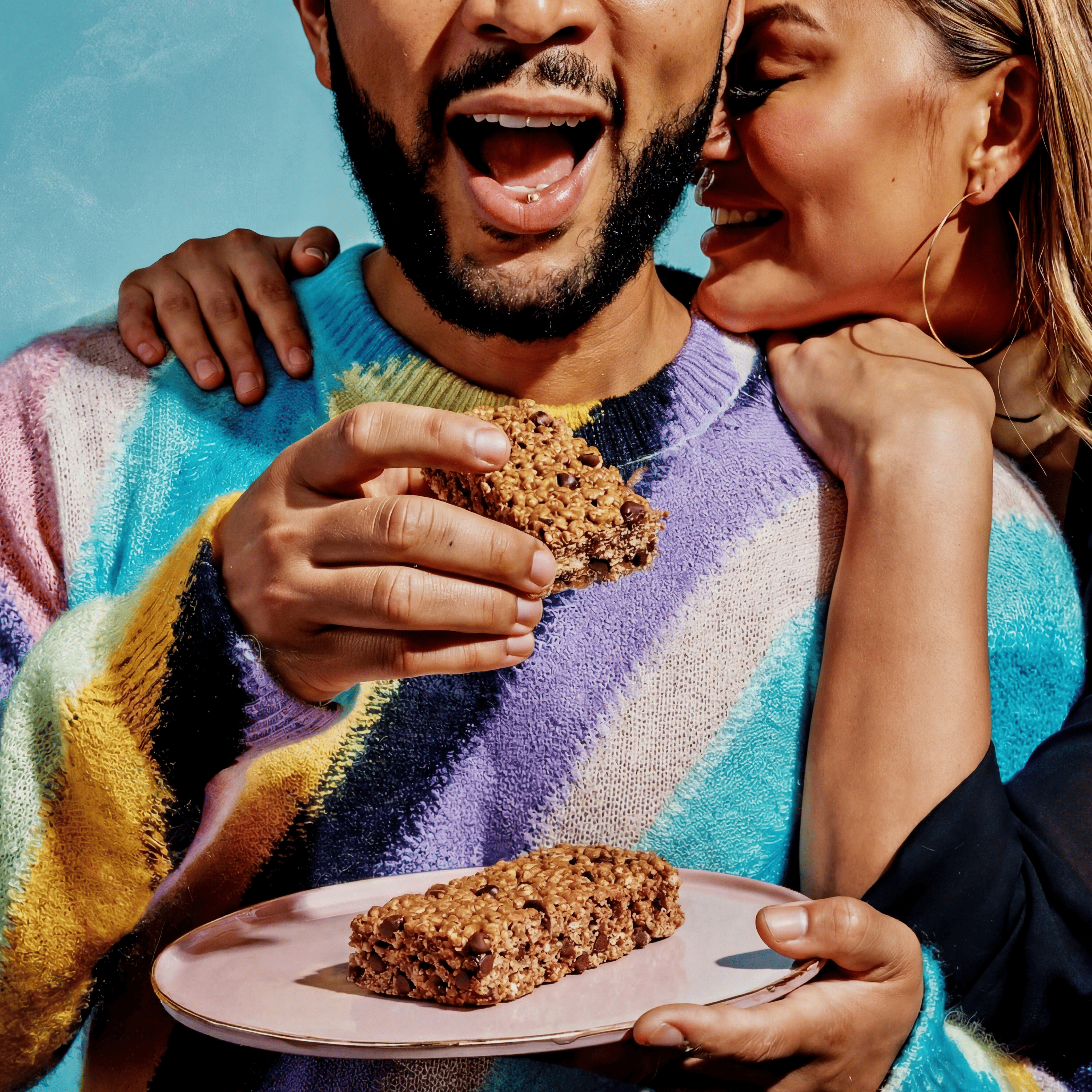 A happy man in a colorful sweater eats a chocolate chip granola bar while a woman hugs him.