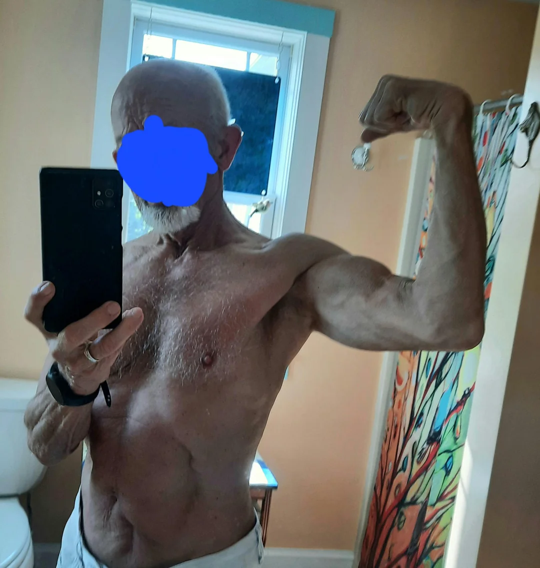 Shirtless man taking a selfie in a bathroom, flexing his arm muscle.