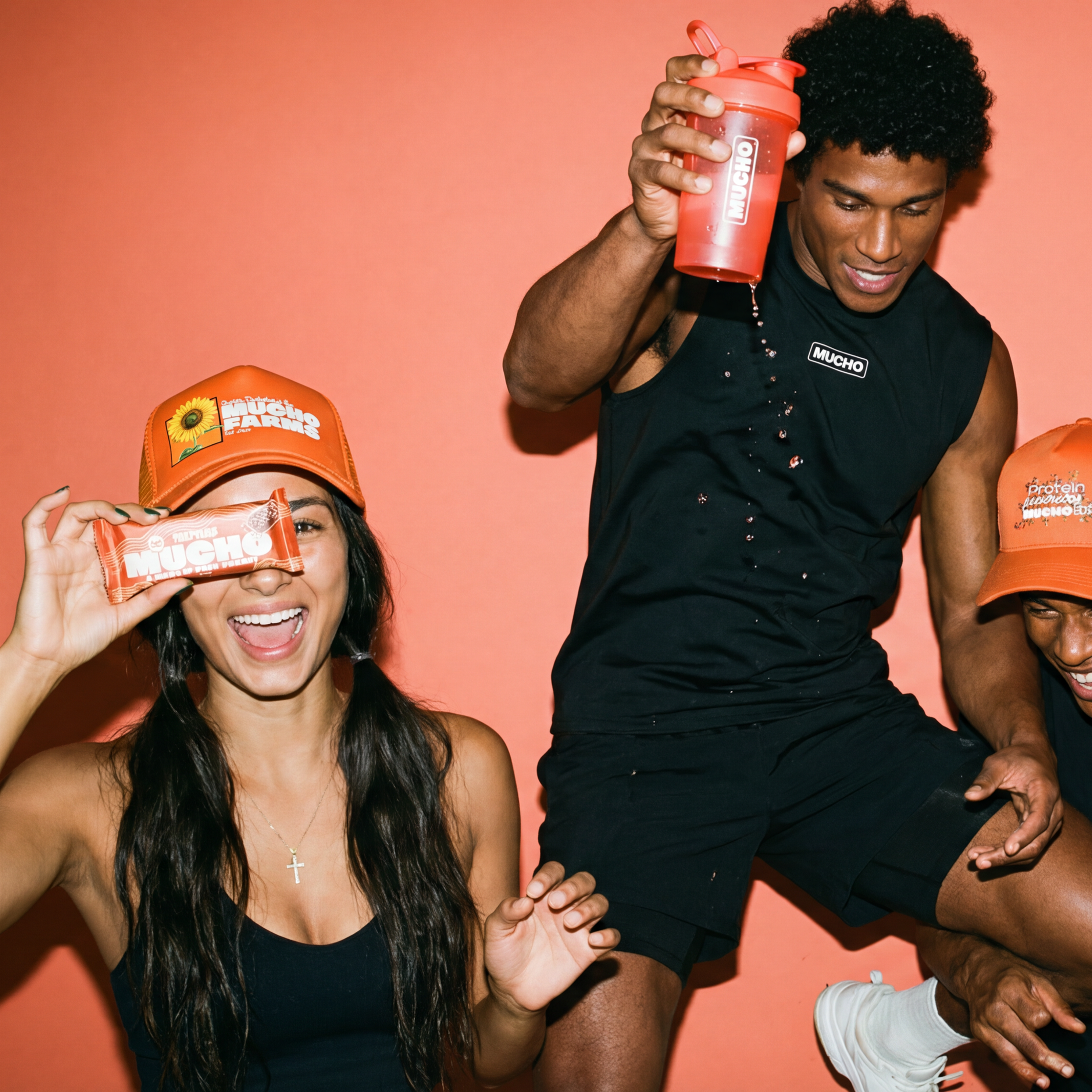Three people playfully pose with Mucho branded hats, a protein bar, and a shaker bottle against an orange backdrop.