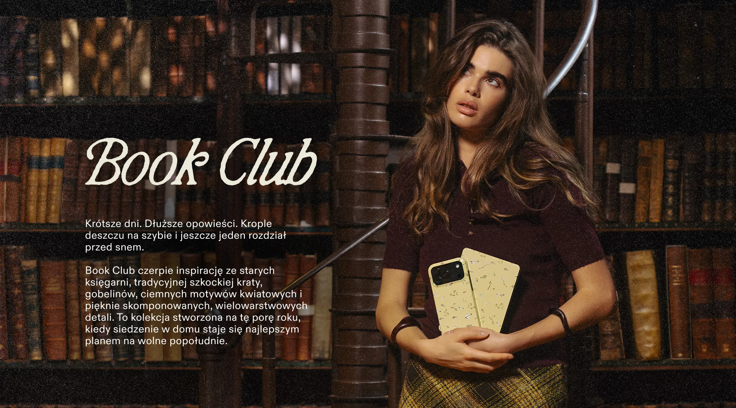 A woman with long brown hair stands in a library, looking up while holding a phone.