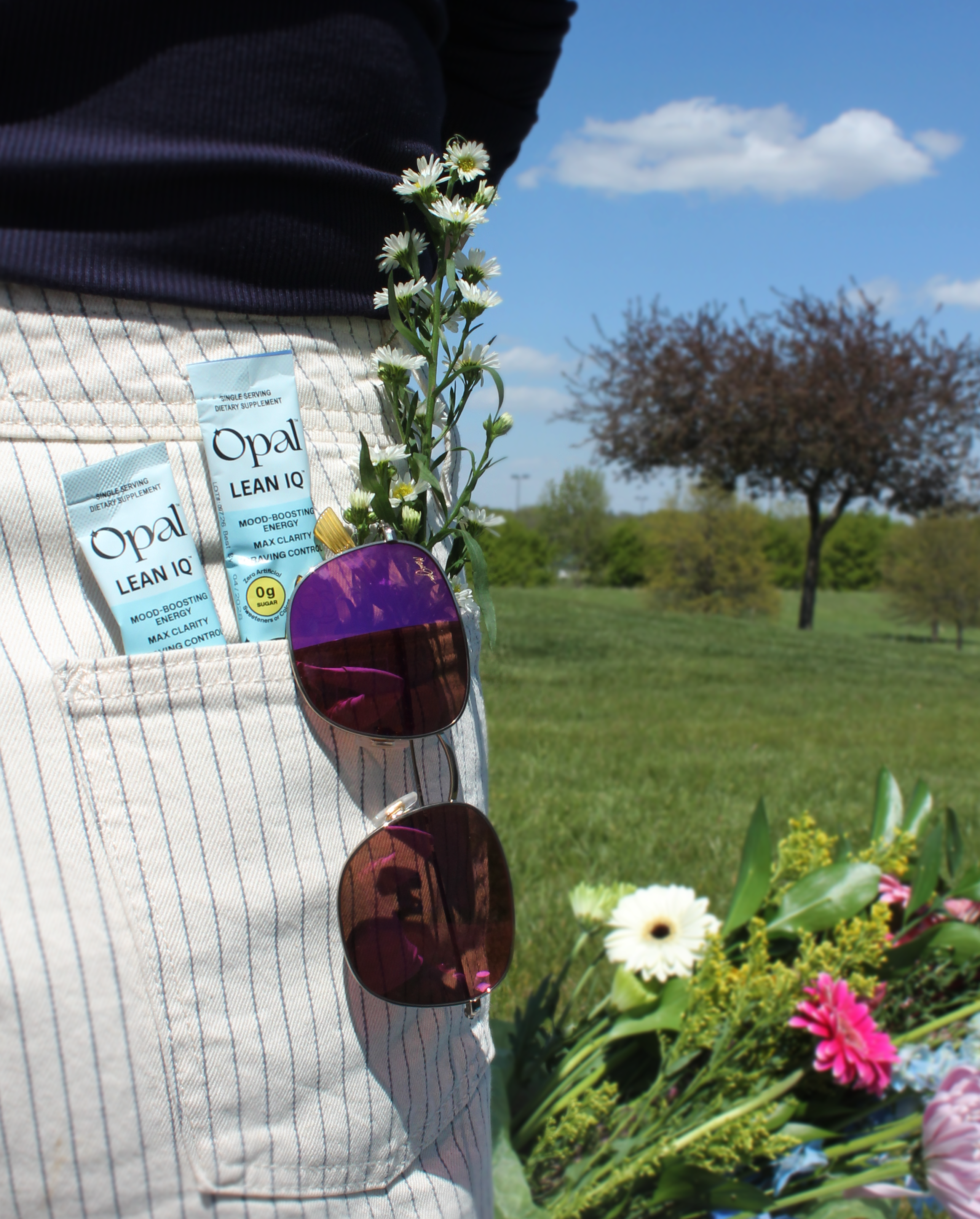 A person's pinstriped pocket holds supplement packets, flowers, and sunglasses, with a park in the background.