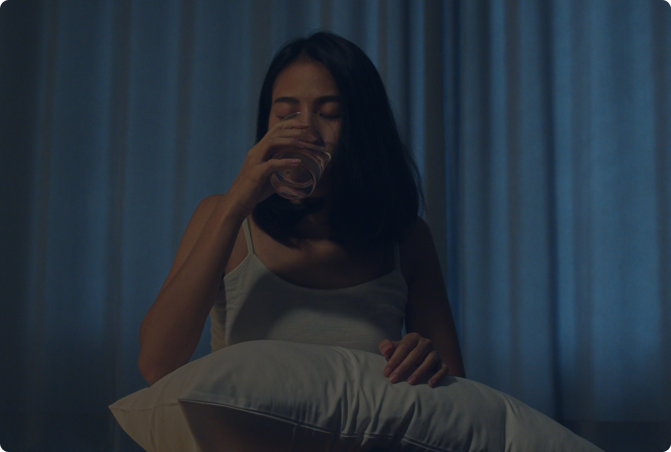A woman in a dimly lit room sits holding a pillow and drinking a glass of water.