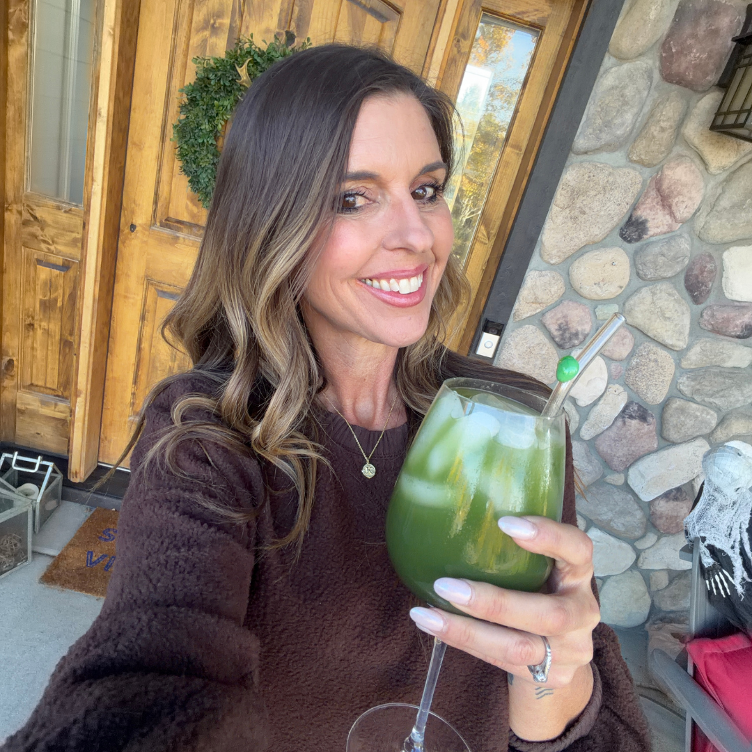 Woman with long hair holding a green drink, standing in front of a wooden door.