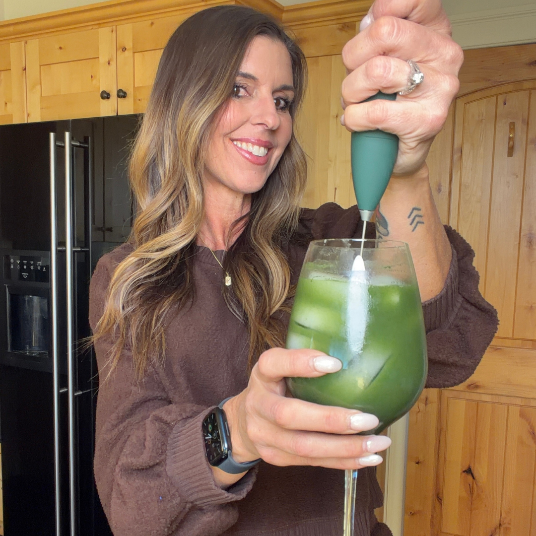 Person blending a green drink in a wine glass in a kitchen setting.