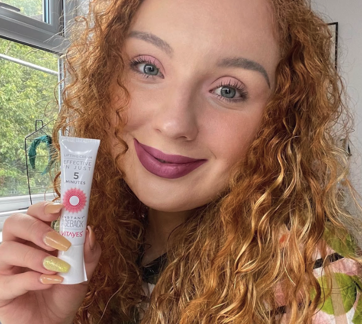 Person with curly hair holding a tube of skincare cream.