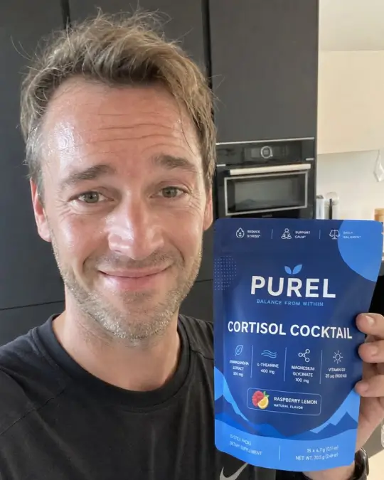A smiling man in a kitchen holds up a blue pouch of PUREL Cortisol Cocktail dietary supplement.
